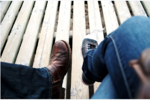 The legs of two people with their legs crossed sitting on a wooden deck.
