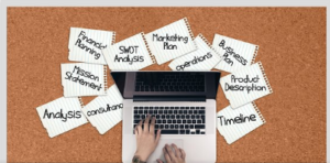 Image of hands on a computer and all components for a business plan around on white paper.