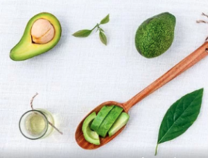 An avocado cut and half put on a wooden spoon beside a glass of oil and another avocado and leaves.
