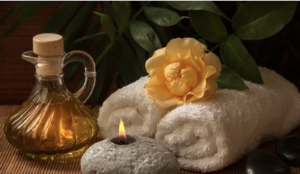 Three white towels with a yellow flower on top and an oil bottle and a lit candle beside them.