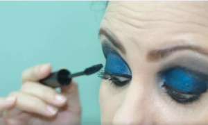 Female with blue eyeshadow applying mascara.