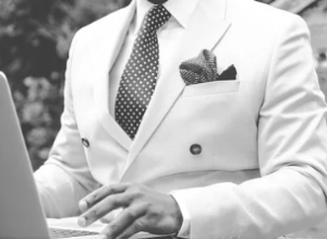 Man formally dressed in a white suite with a grey polka dot tie working on his computer.