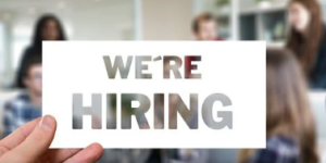 Hand holding a "we're hiring" sign in front of a glass door with people in the background.