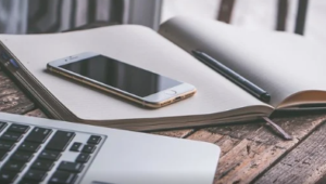 An iphone resting on a open diary with a pen and a computer beside it.