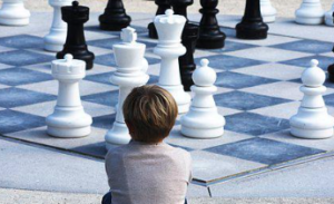 A Person sitting, looking at a life-sized chess board.