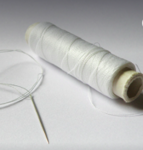 A spool of white thread beside a sewing needle with some thread.