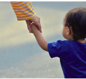 A mother's hand holding a toddler in a blue shirt by hand.