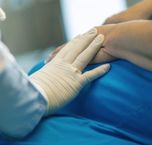 Gloved hand of a practitioner patting hands of patient in a blue gown sitting on a bed.