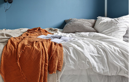 A rumpled bed with messy white sheets and orange blanket in a blue bedroom. 