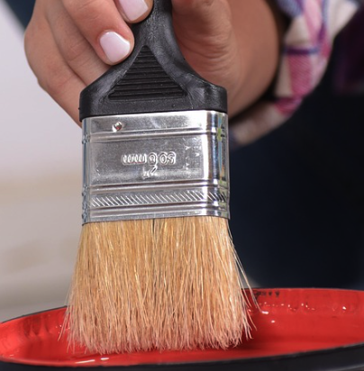 A hand putting a brush in a gallon pan of red paint.